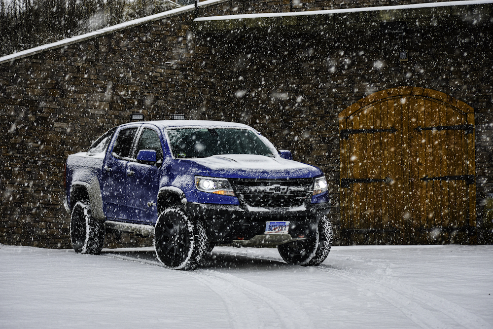 On a snowy day, a blue truck partially covered in snow is parked in front of a medieval style door.