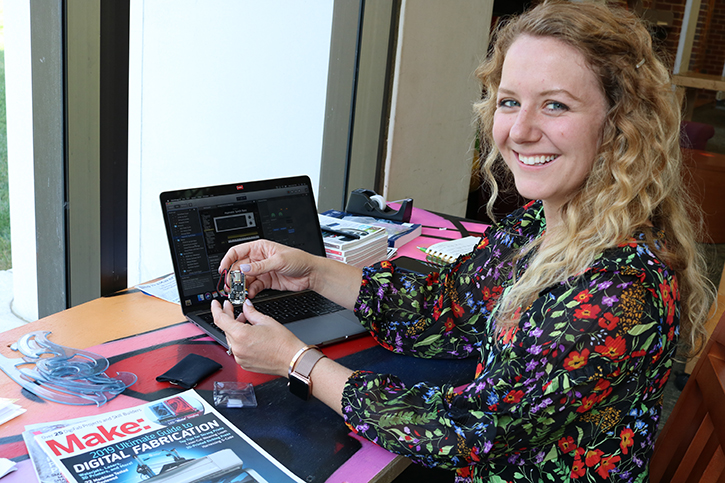 Amanda Davis sits in a chair and turns to smile at the camera. She is holding a wearable electronic prototype device.