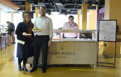 Two people stand in front of a food stand inside Southside dining hall. Another person, looking like they are prepared to serve food, stands behind the card.