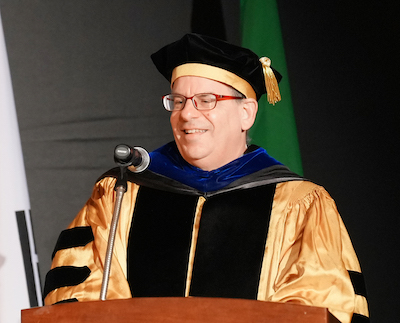 Robert Matz stands at a podium, speaking into a microphone, while wearing full graduation regalia.