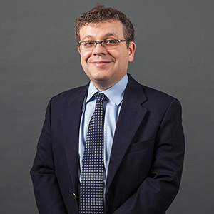 Ilya Somin stands smiling while wearing a black suit and blue collared shirt.