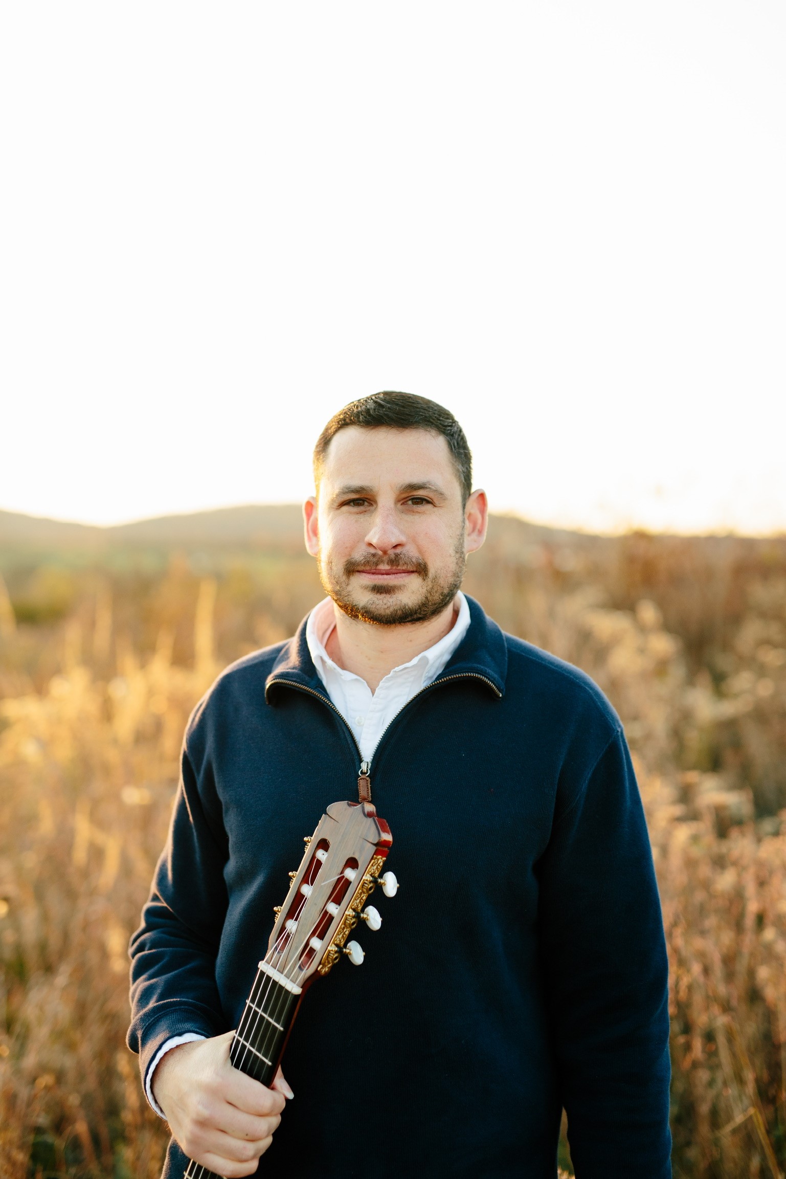 Matt Trkula holding guitar in a field wearing blue sweater