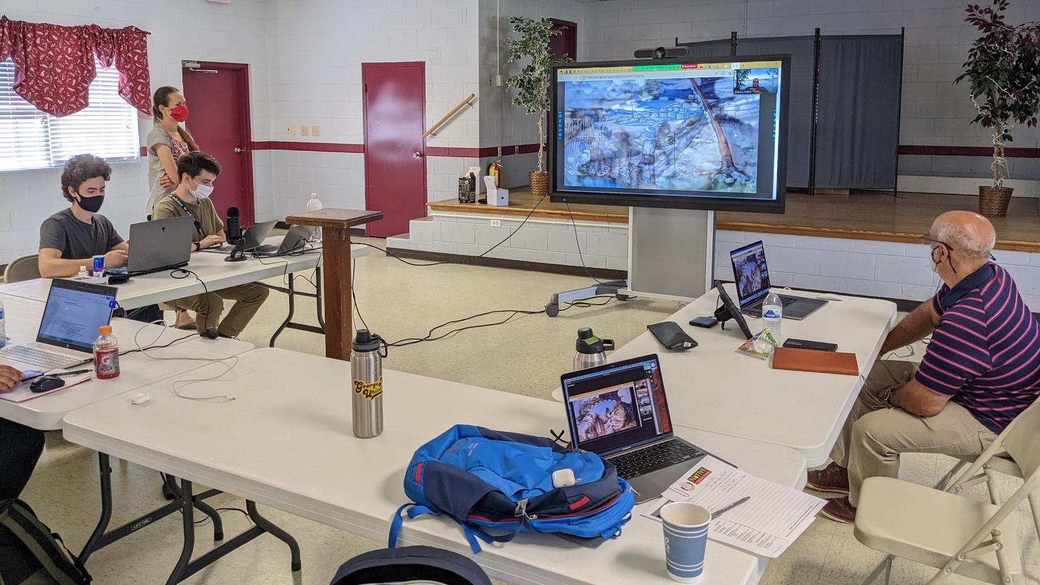 Mason students working with Chickahominy participants in a GIS Skills Building Workshop. They are seating around rectangular folding tables and using laptops. A large screen mirroring one of the laptops is shown up front.