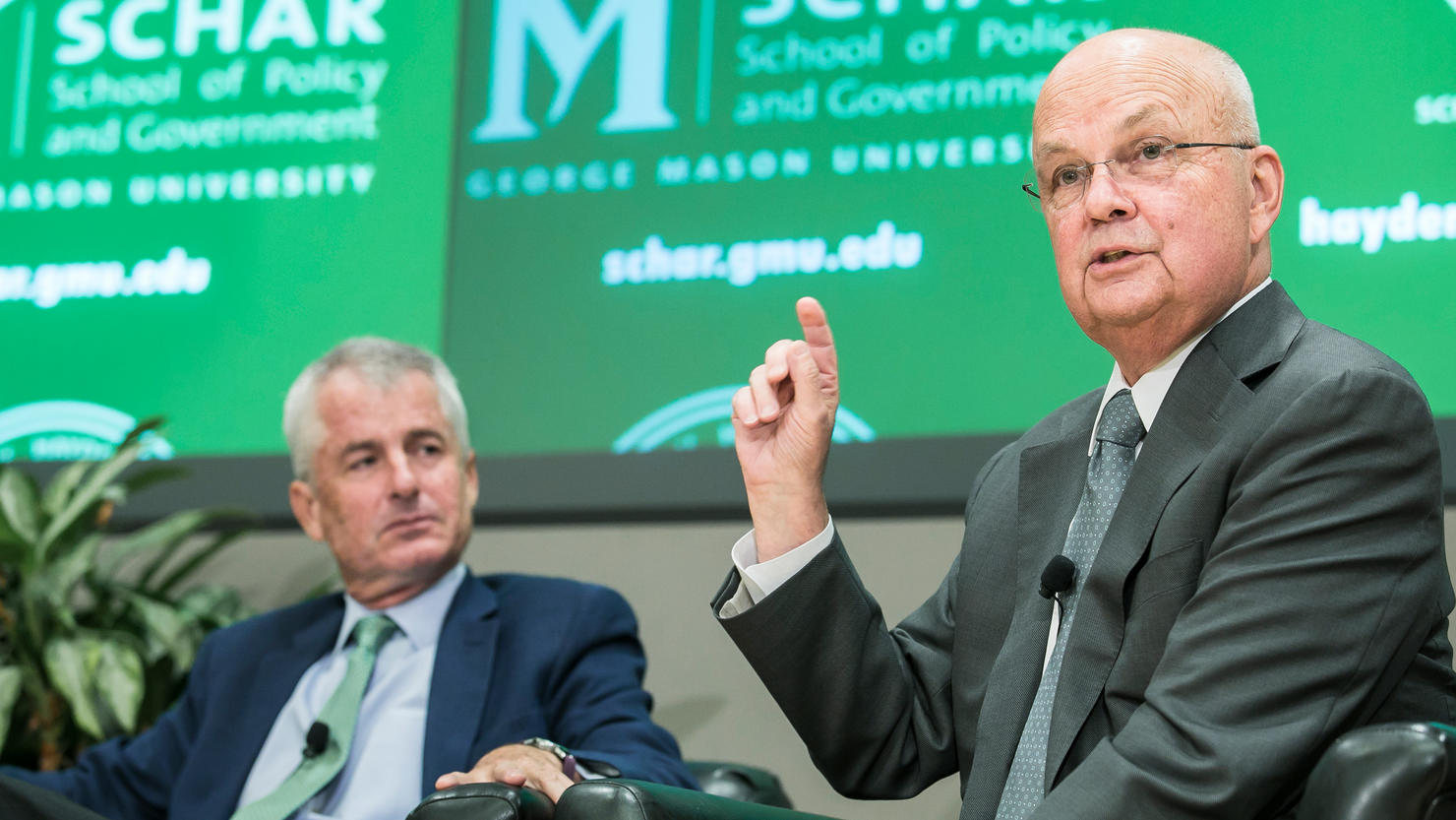 Michael Hayden sitting on a panel. He is speaking with his right hand raised.