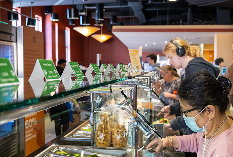 Photo of students getting food at a Mason dining hall. The food station has green card tents on top, labeling the food and its carbon impact