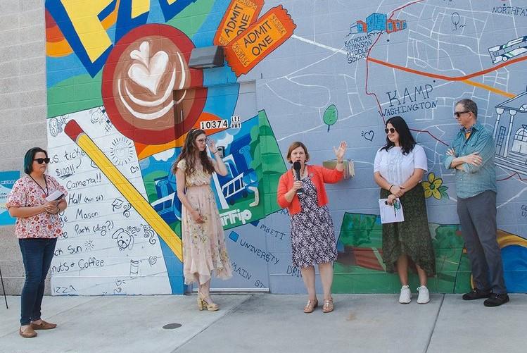 Fairfax City Mayor Catherine Read speaks at the unveiling of the Greetings from Fairfax mural. She is holding a microphone and next to her is artist Lecsi Pillar