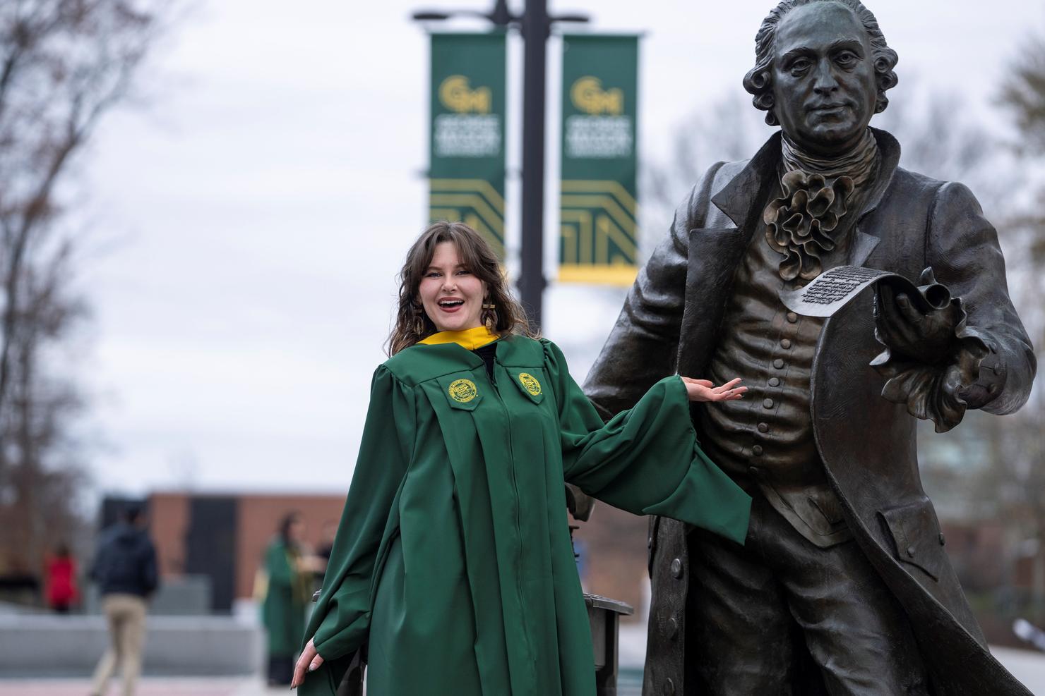 A graduate wearing regalia poses in celebration at the George Mason statue on Wilkins Plaza