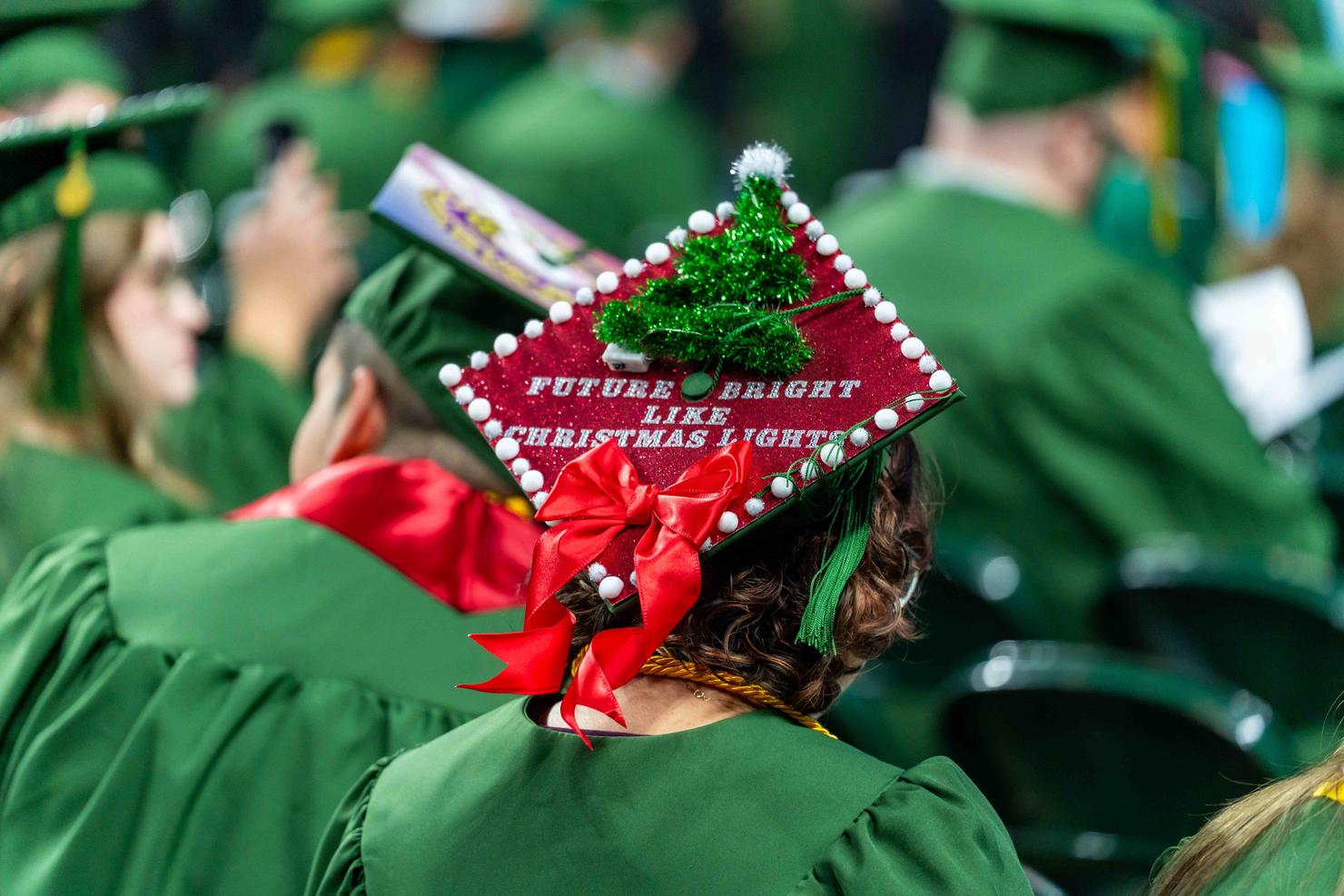 A decorated graduation cap in the crowd has a green tinsel tree and says "Future bright like Christmas lights"