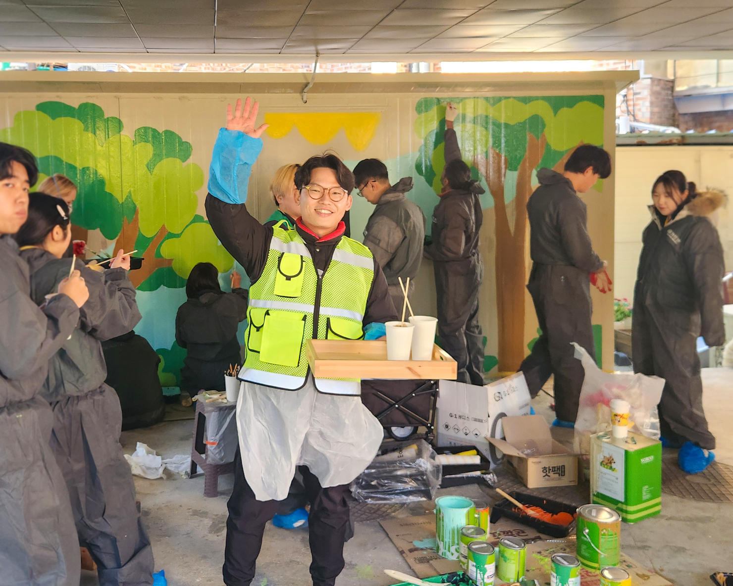 Volunteer painter wearing protective gear holds up a tray of paint, and smiles and waves