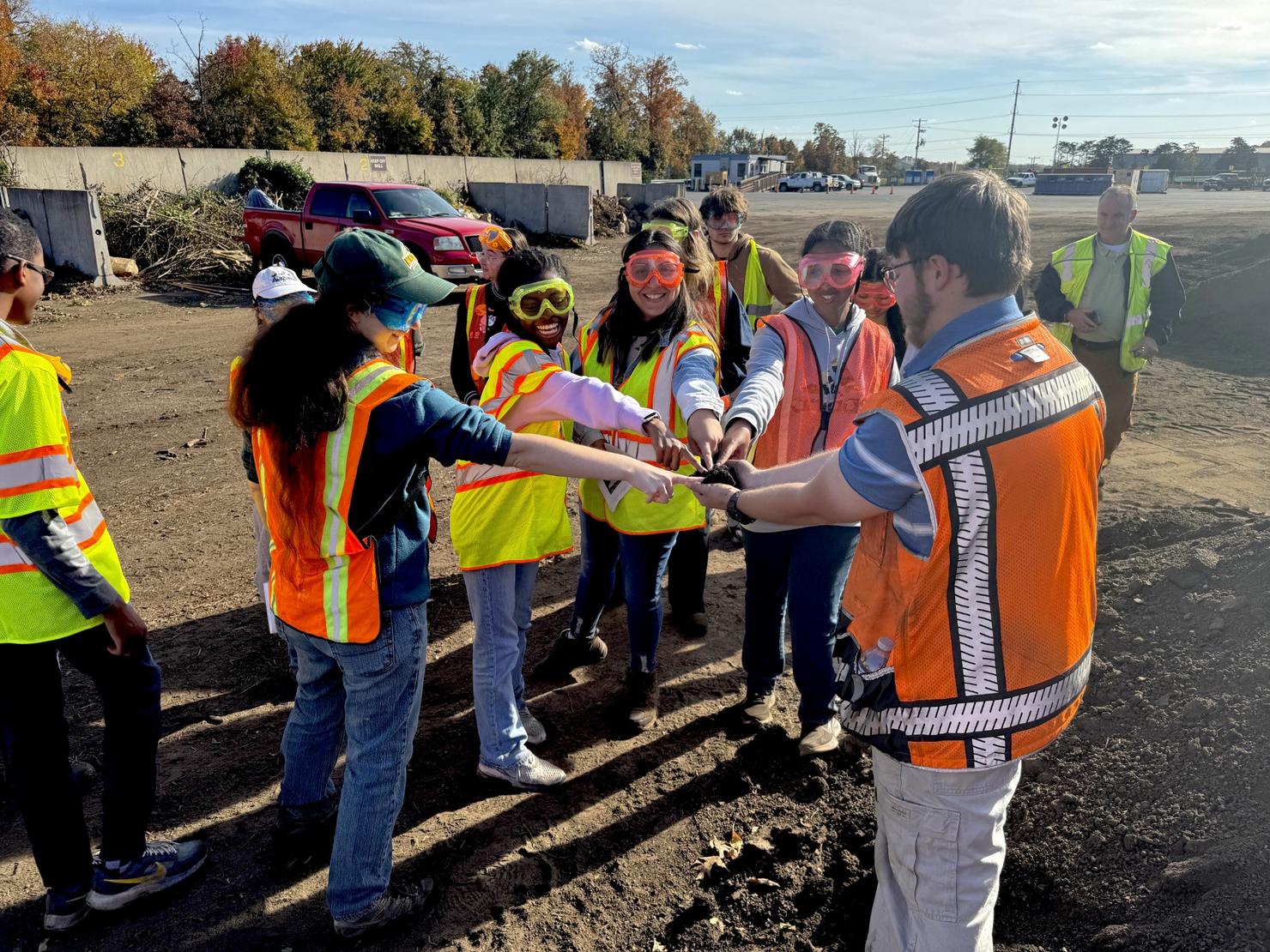 Students gather for a group high-five during a field trip to the composting facility. They are wearing goggles and reflective vests