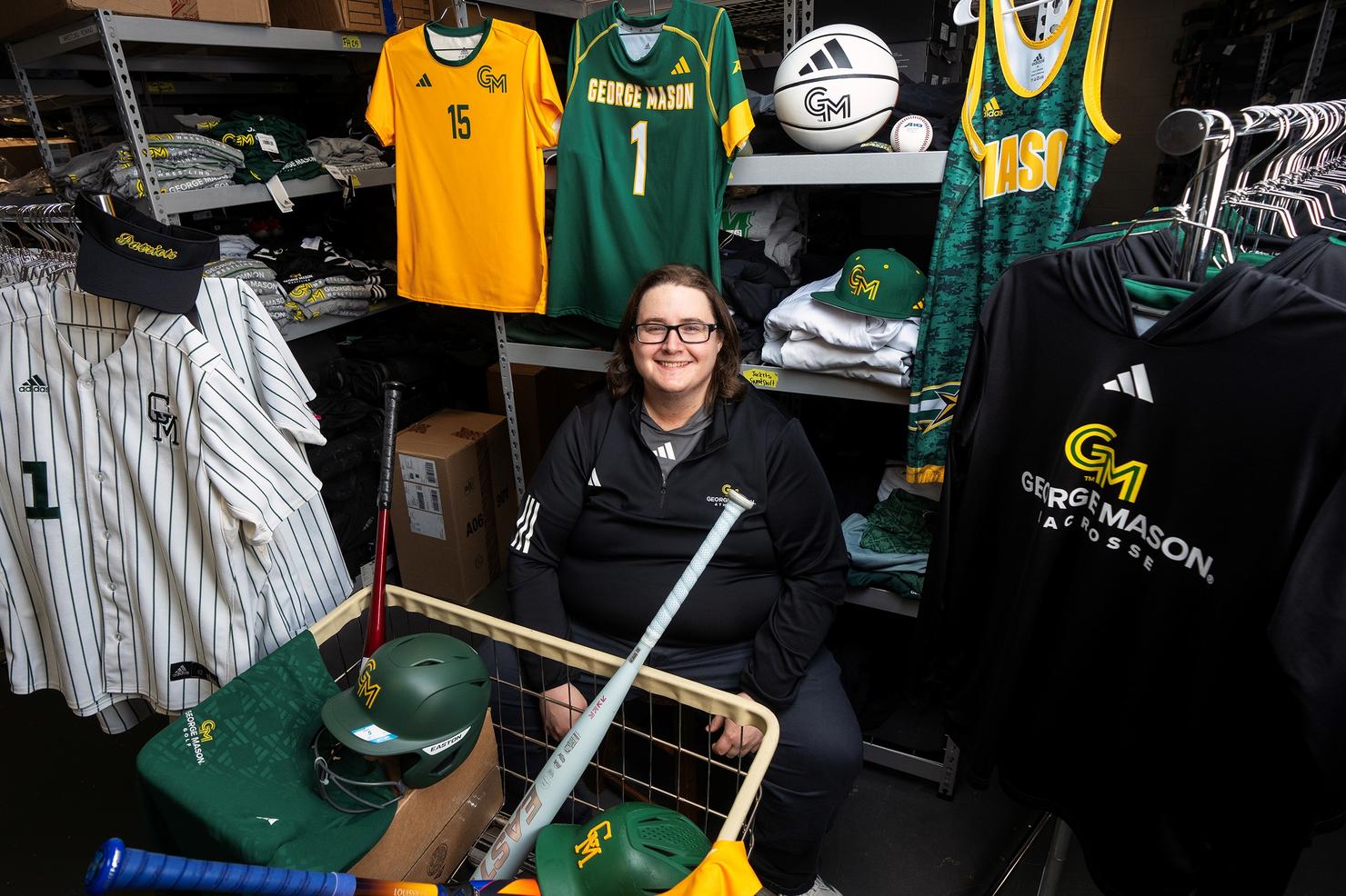 CJ MInteer sits in a storage room, surrounded by George Mason Athletics equipment and jerseys