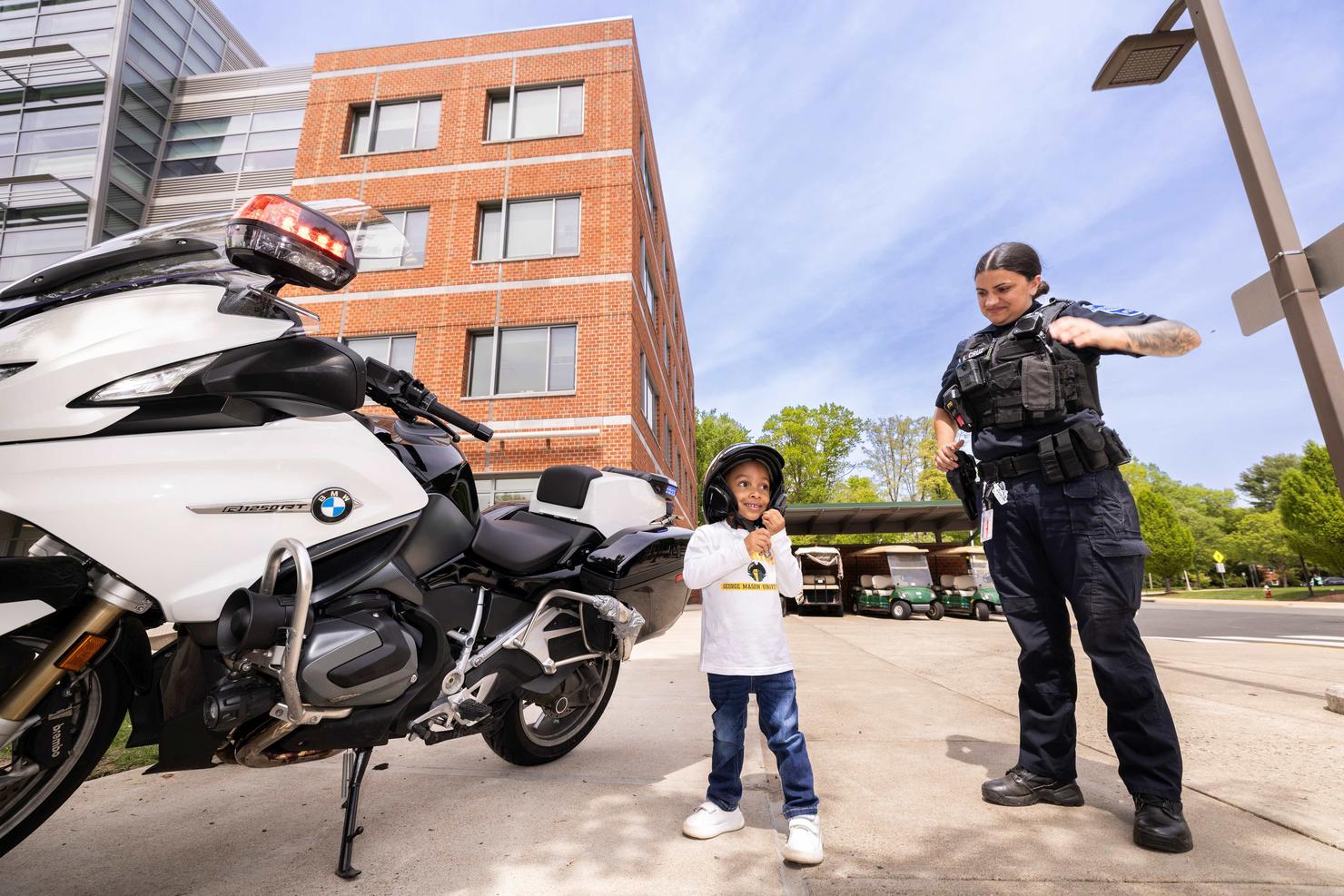 A young child tries on a police motorcycle helmet next to the police motorcycle, with the officer standing by