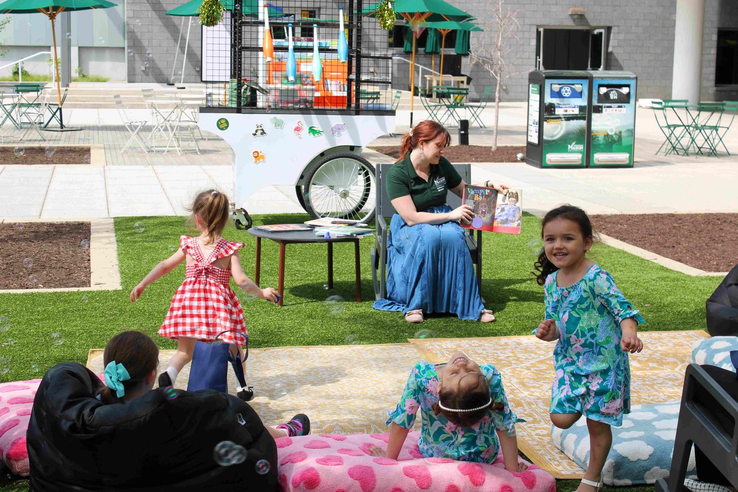 An adult reads a picture book to children on the plaza. The kids are moving around the area