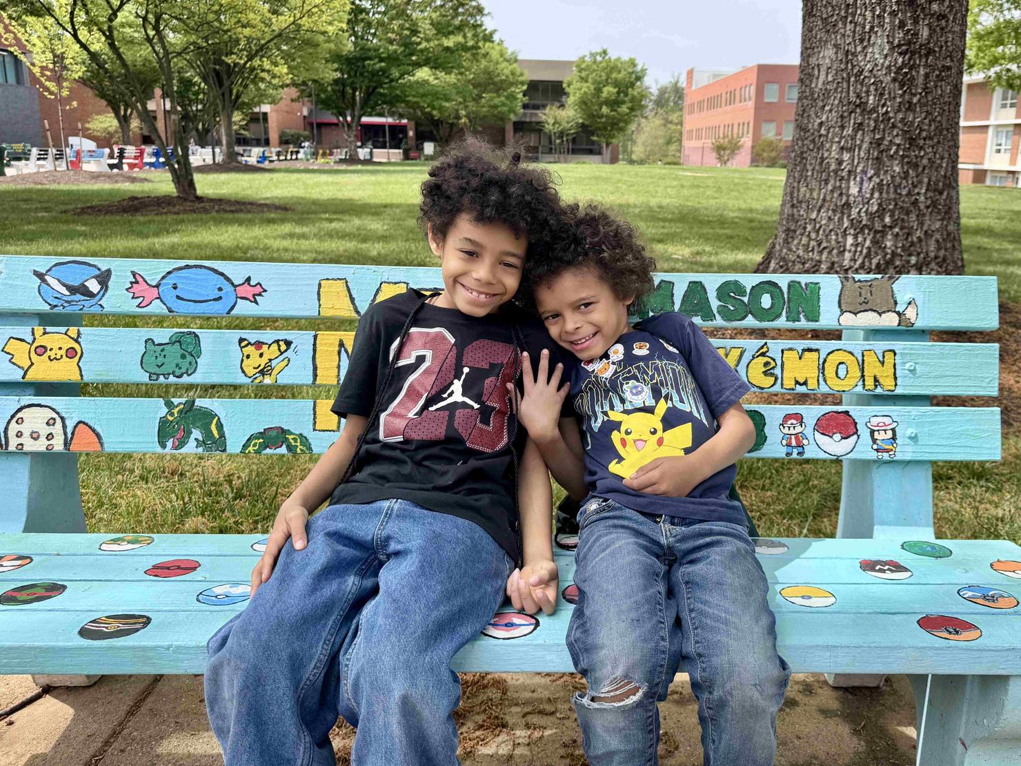 Two children sit together smiling on the bench decorated by the George Mason Pokemon Club