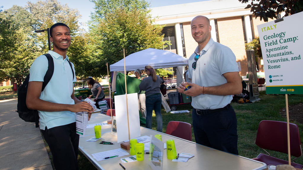 Student talks with a study abroad representative at the Study Abroad fair on the quad outside the Fenwick Library. A sign promotes a trip to Italy for a Geology Field Camp.