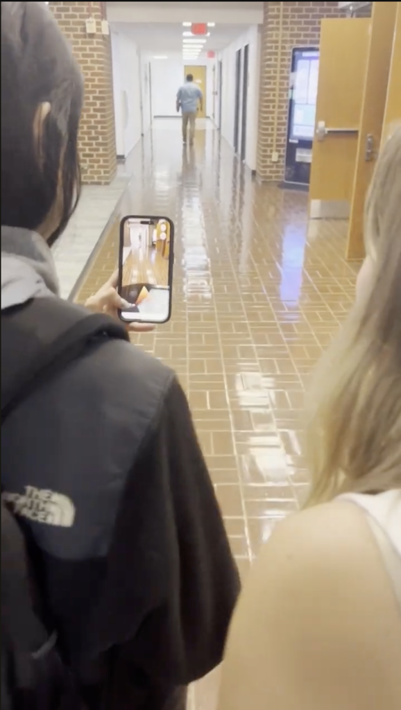 Two students look at a phone before them, which displays an augmented reality view of directions through the hallway before them.