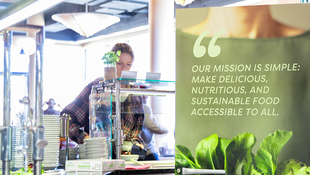 A student serves themself food from a buffet. Visible on a wall is the quote "Our mission is simple: make delicious, nutritious, and sustainable food accessible to all."