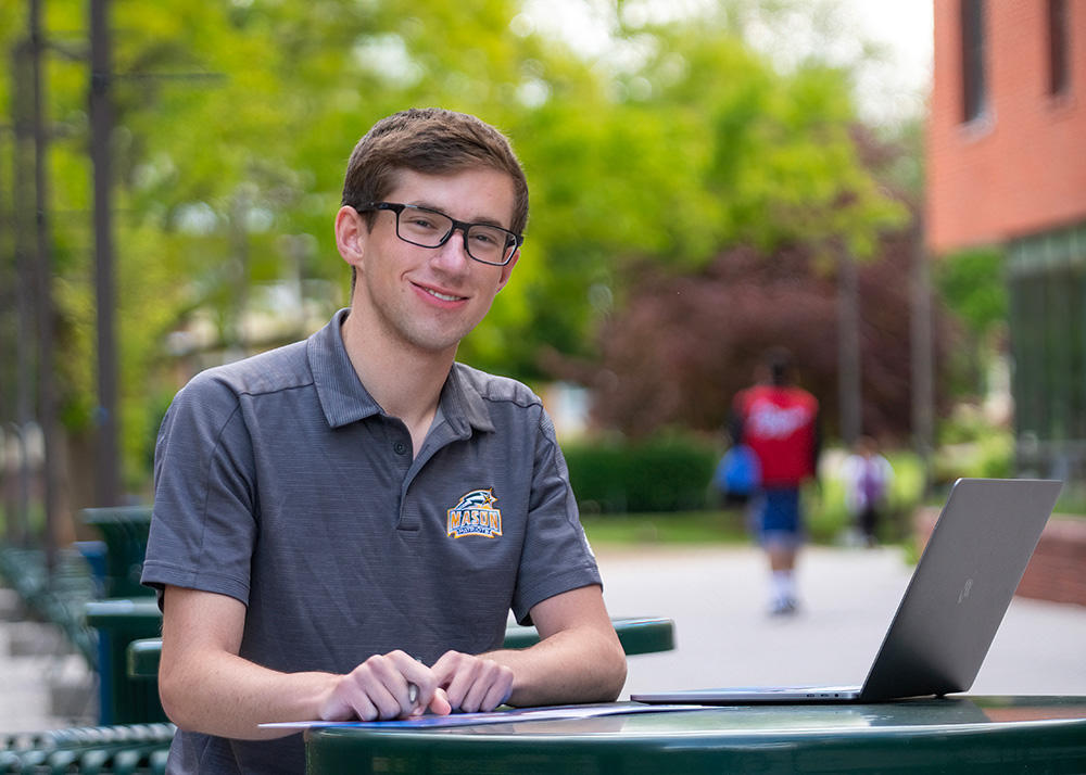 A student sits working on a laptop at a picnic table on campus.