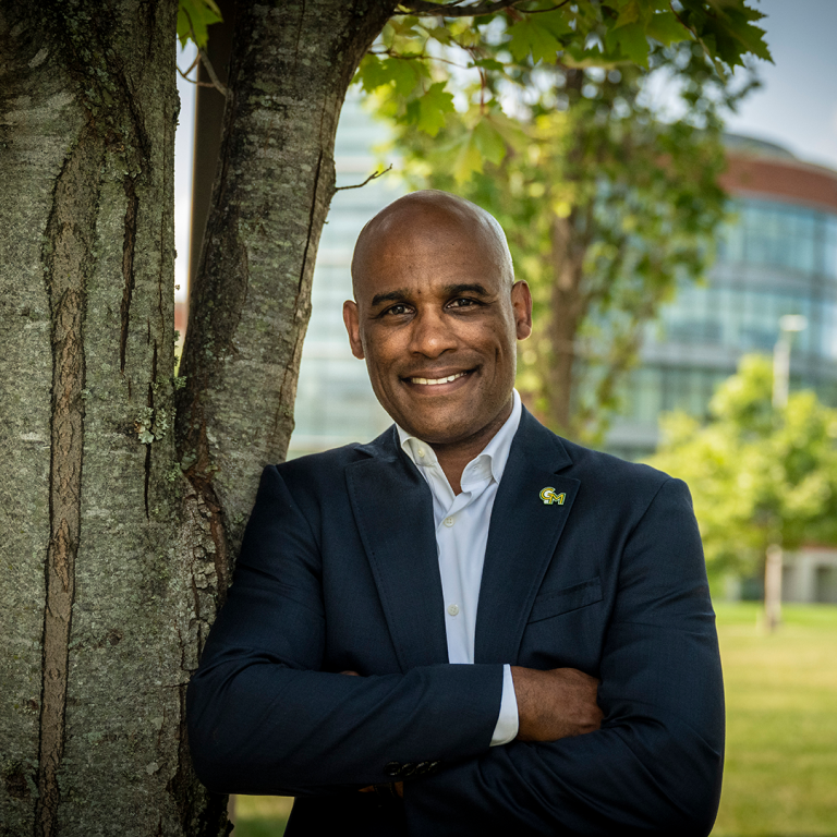 A Black man wearing a blue blazer and white button down open collared shirt, leans against a tree on campus on a sunny day.