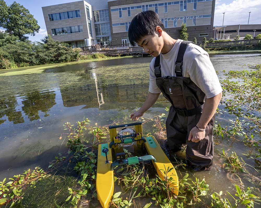 A student in full body wet/rubber suit stands in thigh high water with an autonomous boat that collects water samples