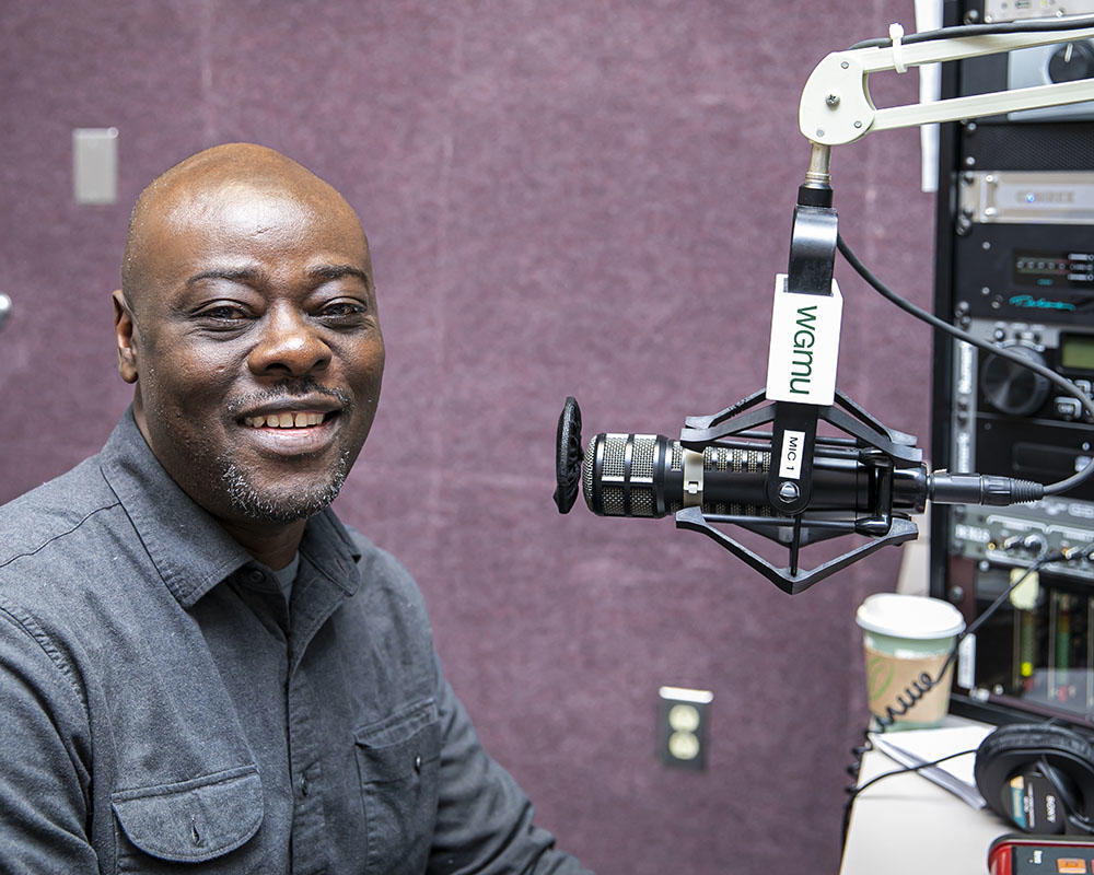 A Black man wearing a blue button down shirt with breast pockets speaks into a microphone