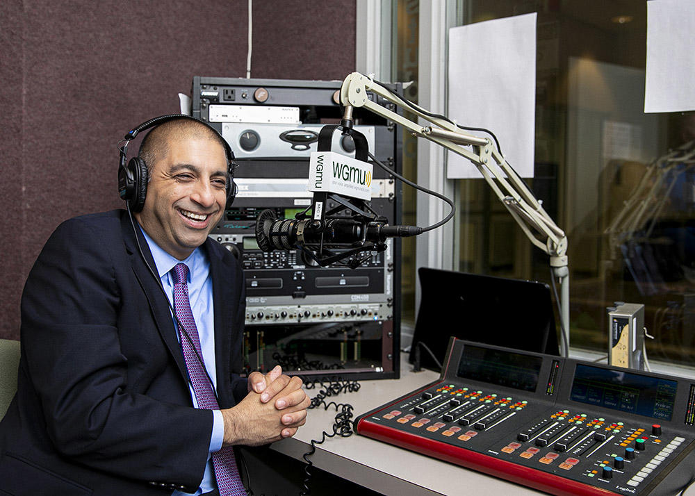 A man wearing a blazer, tie and button-down shirt, wears headphones as he speaks into a microphone in a recording booth.