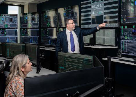 A woman sitting at a computer, while a man points to a large mainframe computer.