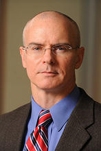 Koper wears a dark sportcoat, blue button up shirt and a red tie with blue and white diagonal stripes