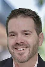 Shane Caswell is wearing a white collared shirt and dark blazer