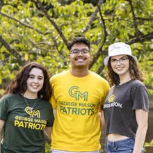 Three students pose together in front of a tree while wearing George Mason University attire.
