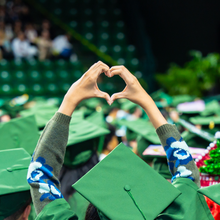 A graduate in a sea of other graduates has their hands raised into a heart over their head.