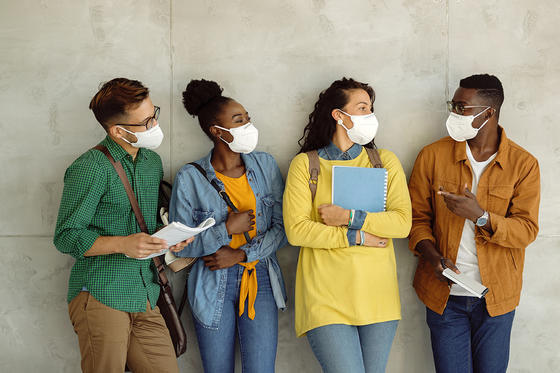 a group of four multiethnic students standing against a wall all wearing protective masks