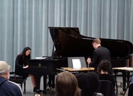 Catherine "Kitty" Fields, a young woman with long dark hair wearing a dark, floor length dress, is seated at a grand piano performing her work, accompanied by Hunter Wirt, a young white man with light brown hair who is seen in the foreground of the photo with his back to the camera.