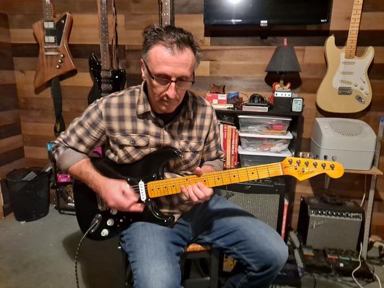 Bob Vay sits and plays a black electric guitar. A wooden paneled wall behind him has several other electric guitars hanging.