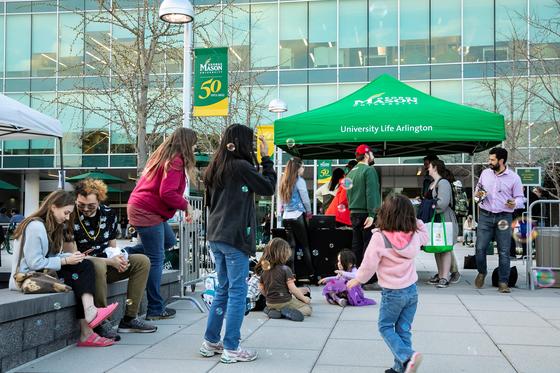 Photo of a group of adults and children gathered on the plaza at Mason Square, with small green tent that says University Life. The children are blowing bubbles and playing