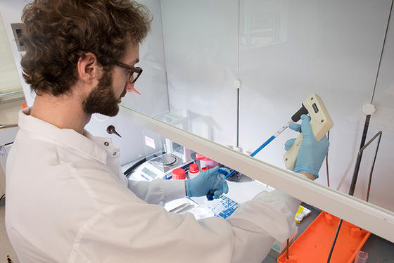 A man in a lab coat and gloves works with a syringe and test tubes in a lab environment.