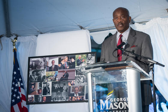 Colbert King delivers a speech from the podium at the dedication of Roger Wilkins Plaza. He wears a suit and tie, and is flanked by the american flag and a tableau of photographs memorializing Wilkins