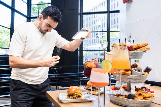 Alex Hussein stands positioning a light in one hand and a camera phone in the other, preparing to take a photo of the spread of burgers, appetizers, and drinks in front of him.