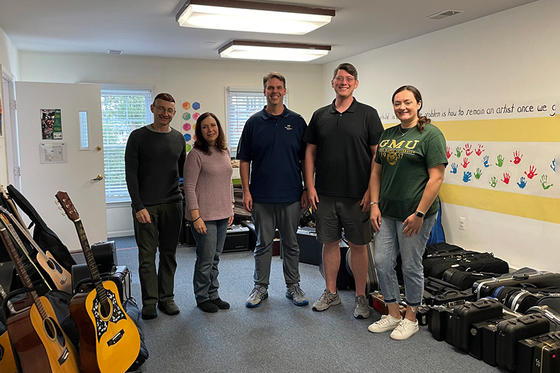 Five adults stand in what looks to be a childrens' classroom, surrounded by multiple guitars and many other instruments in cases.
