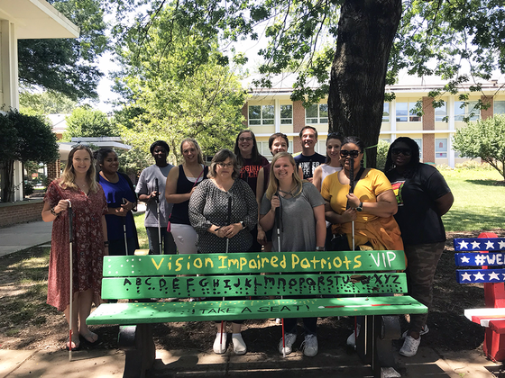 Students with sight canes stand around a green bench painted to say "Vision Impaired Patriots" and a translation of the braille alphabet