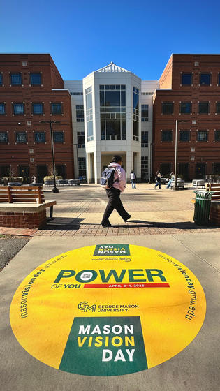 A student walks near the Johnson Center. On the ground is visible a decal advertising Mason Vision Day.