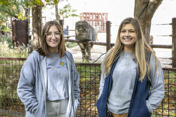 Smithsonian-Mason School of Conservation student Eva Noroski and Mason alumna and Elephant Trails keeper Ashley Fortner stand side-by-side in front of an elephant enclosure at the National Zoo. A small Asian elephant resting her trunk on the enclosure railing peeks in between the two women.