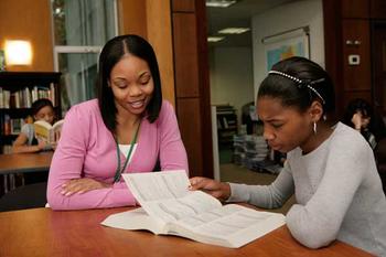 A woman and a girl sitting at a library table and looking at a textbook.