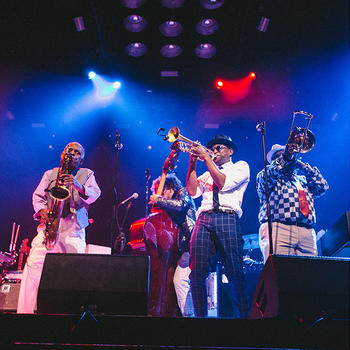 Members of the Preservation Hall Jazz Band perform onstage behind audio speakers, playing saxophone, upright bass, trumpet, and trombone.