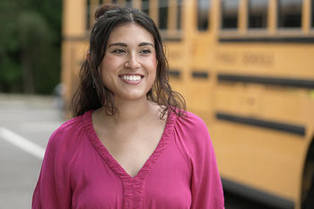 A woman looks into the distance, smiling. Behind her is a school bus.