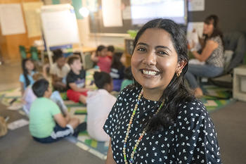 A woman poses for the classroom. Behind her is a class of young students, sitting on the floor, listening to another teacher telling them a story.
