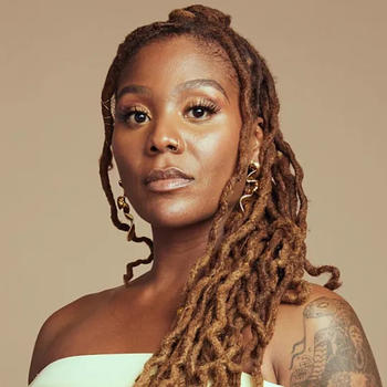 A Black woman looks at the camera. She has long braided hair, a strapless white dress, and a tatoo on her left shoulder.