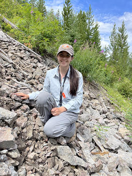 Mateo kneels on a scree field in Yellowstone Park, green foliage behind her. She smiles for the camera.