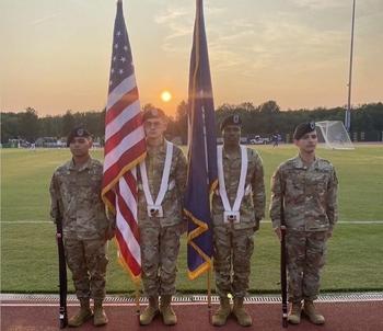 Four ROTC candidates, two holding flags, stand at attention for a flag ceremony as the sun sets behind them.