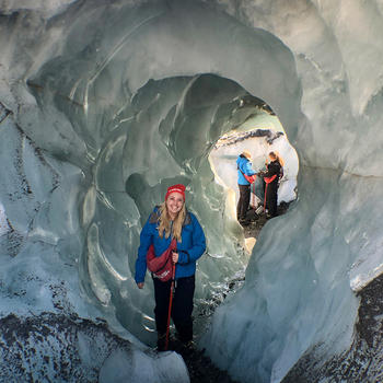 A student in winter attire stands in a round tunnel through a glacier. Two more people can be seen behind them.